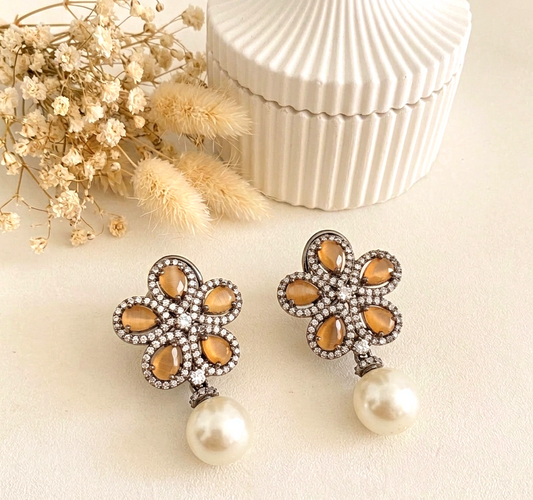 A pair of floral drop earrings with amber-colored teardrop stones, a halo of clear rhinestones, and a dangling white pearl, displayed on a light cream surface with dried flowers and a white ribbed box in the background.