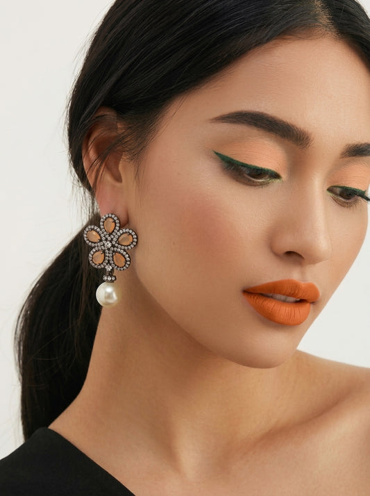 A close-up of a woman wearing a large, ornate flower-shaped dangle earring with a white pearl drop. The earring features a silver-toned metal setting with small clear stones and peach-colored petal inlays.