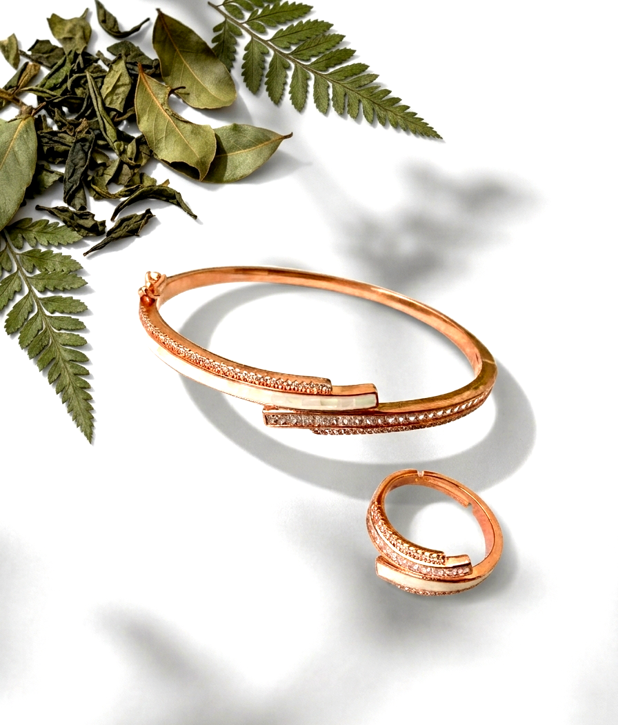 A rose gold bangle bracelet and matching ring with pavé-set clear stones, styled on a white surface with dried green leaves and ferns.