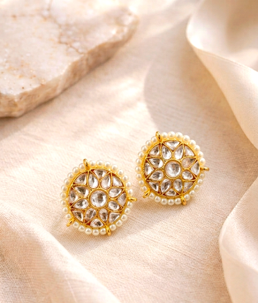 A pair of gold-toned circular stud earrings with clear stones and a border of small white pearls, displayed on draped cream fabric with a stone prop in the background.