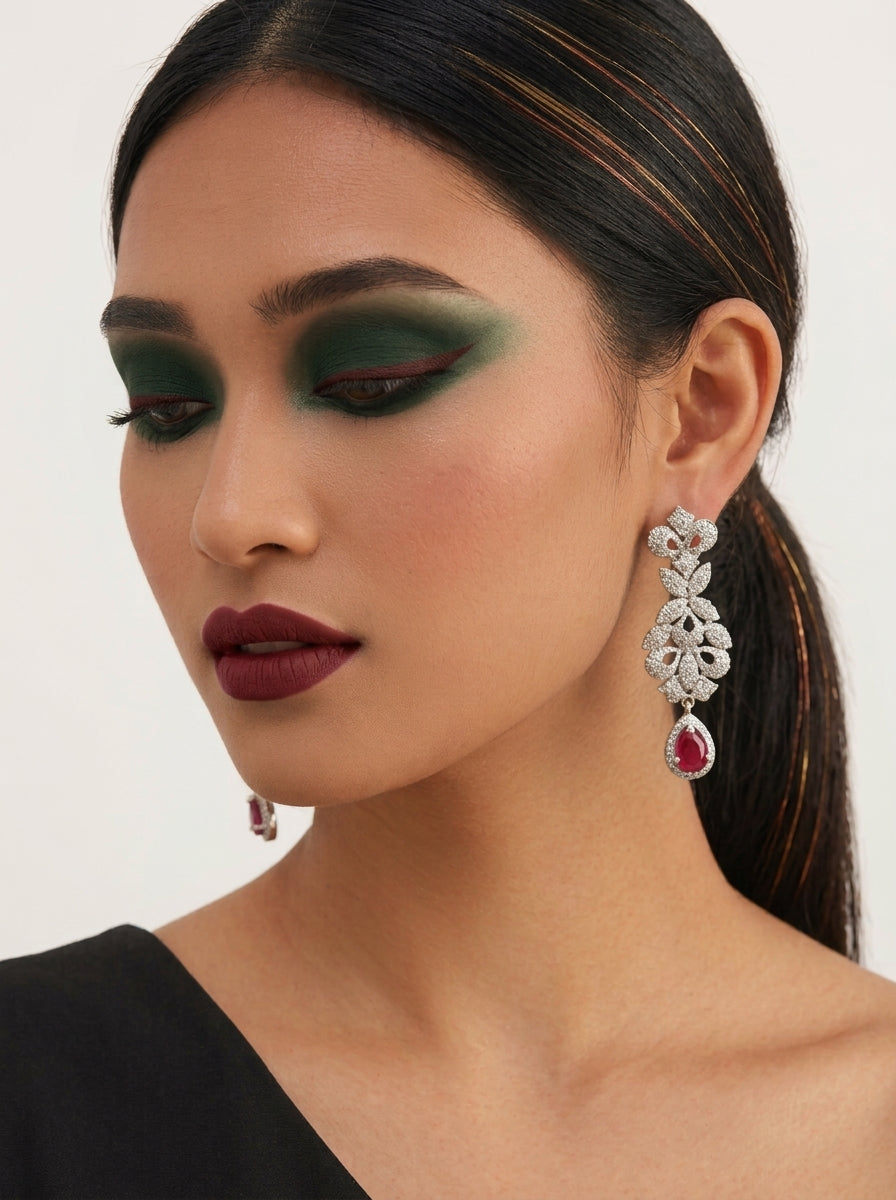 A close-up of a model wearing a silver-toned, multi-tiered drop earring with a red teardrop gemstone and clear stones, styled with dark green eyeshadow and dark red lipstick.