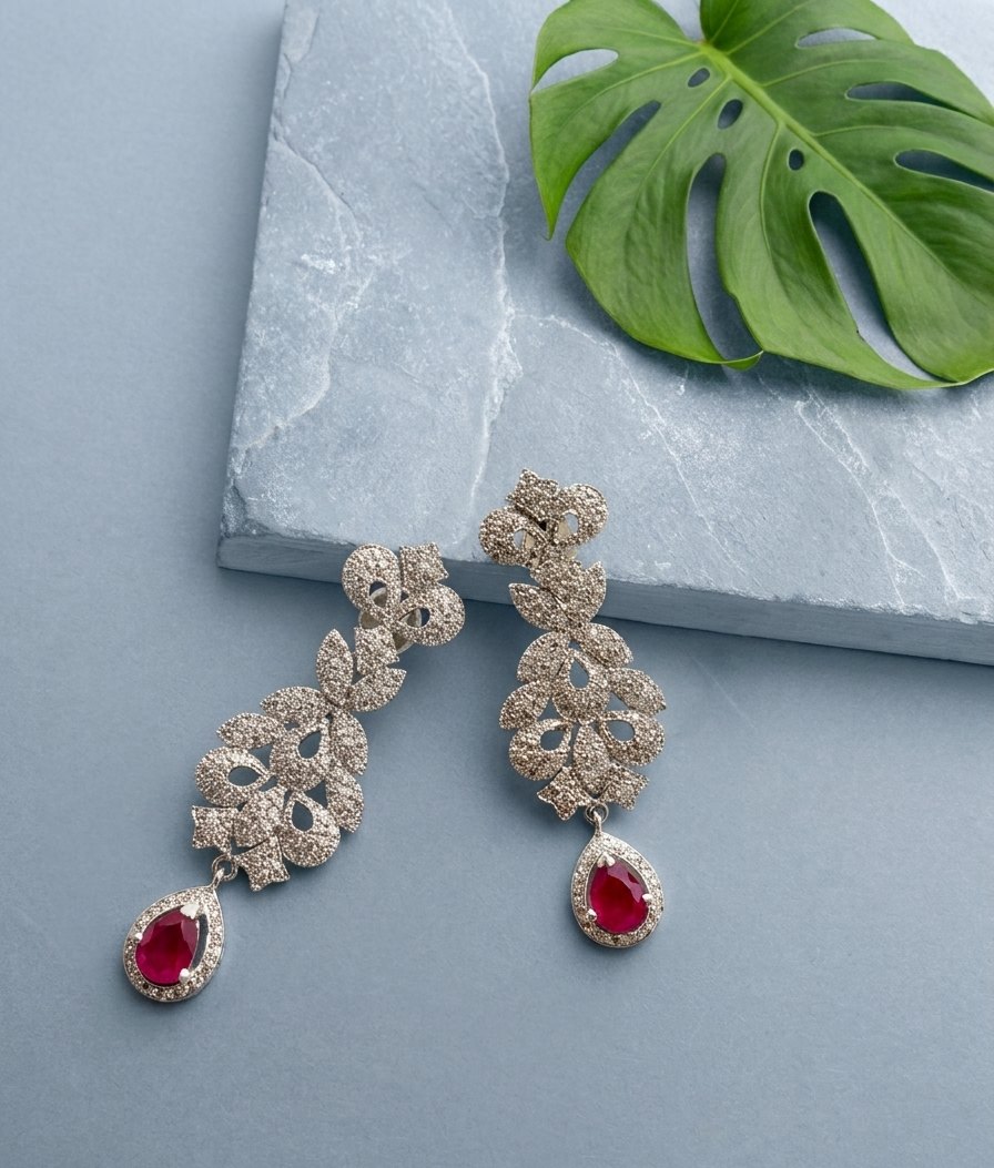 A pair of gold-toned dangle earrings with floral clusters of clear stones and teardrop-shaped red gemstones, displayed on a light blue-gray surface next to a green monstera leaf and a gray stone slab.