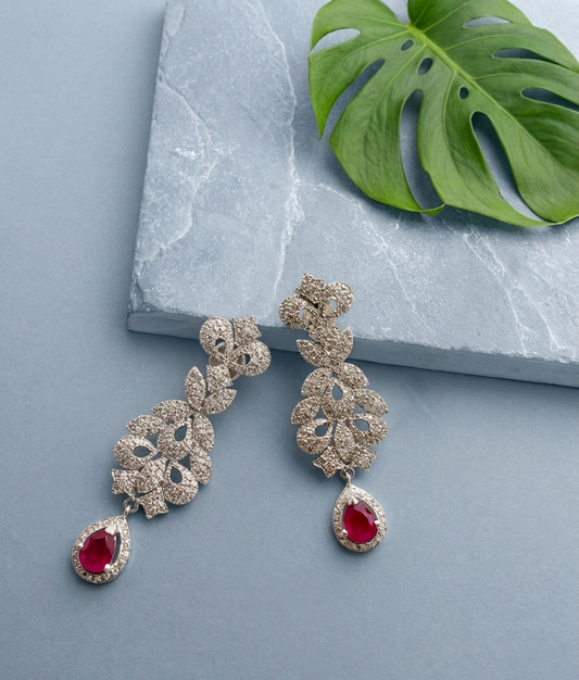 A pair of gold-toned dangle earrings with floral clusters of clear stones and teardrop-shaped red gemstones, displayed on a light blue-gray surface next to a green monstera leaf and a gray stone slab.