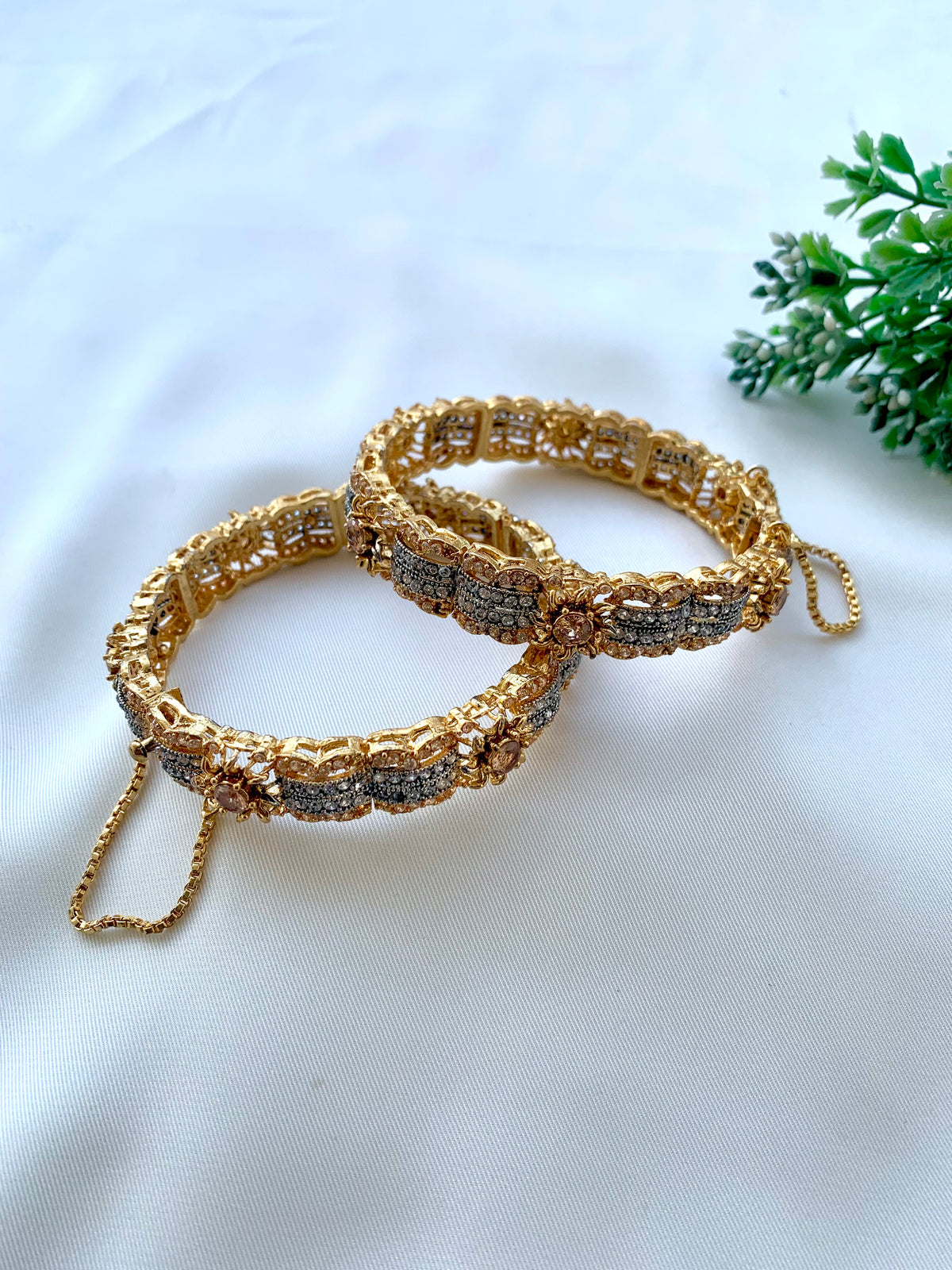 Two ornate gold-toned bangles with silver-toned beaded detailing, displayed on a white fabric background with a small green plant in the corner.