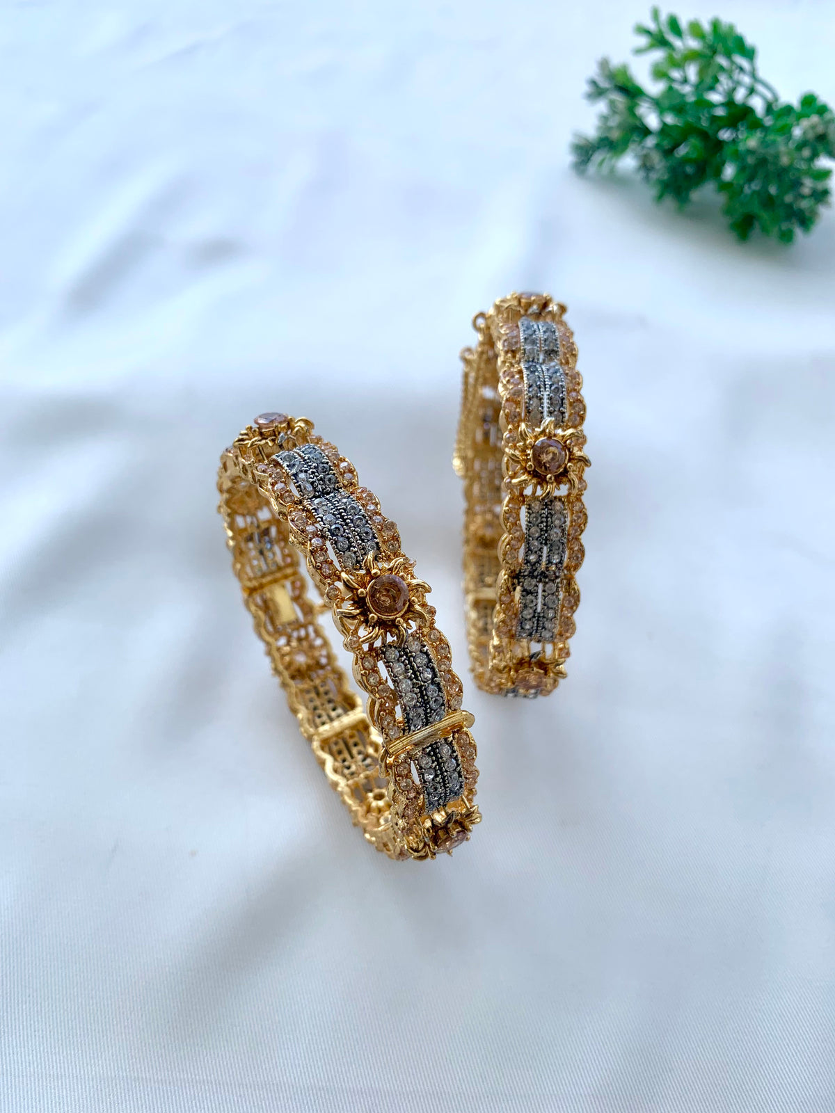 A pair of gold-plated bangles decorated with silver-toned pavé stones and small rose-gold center stones, displayed on a white fabric background with a small green plant in the corner.