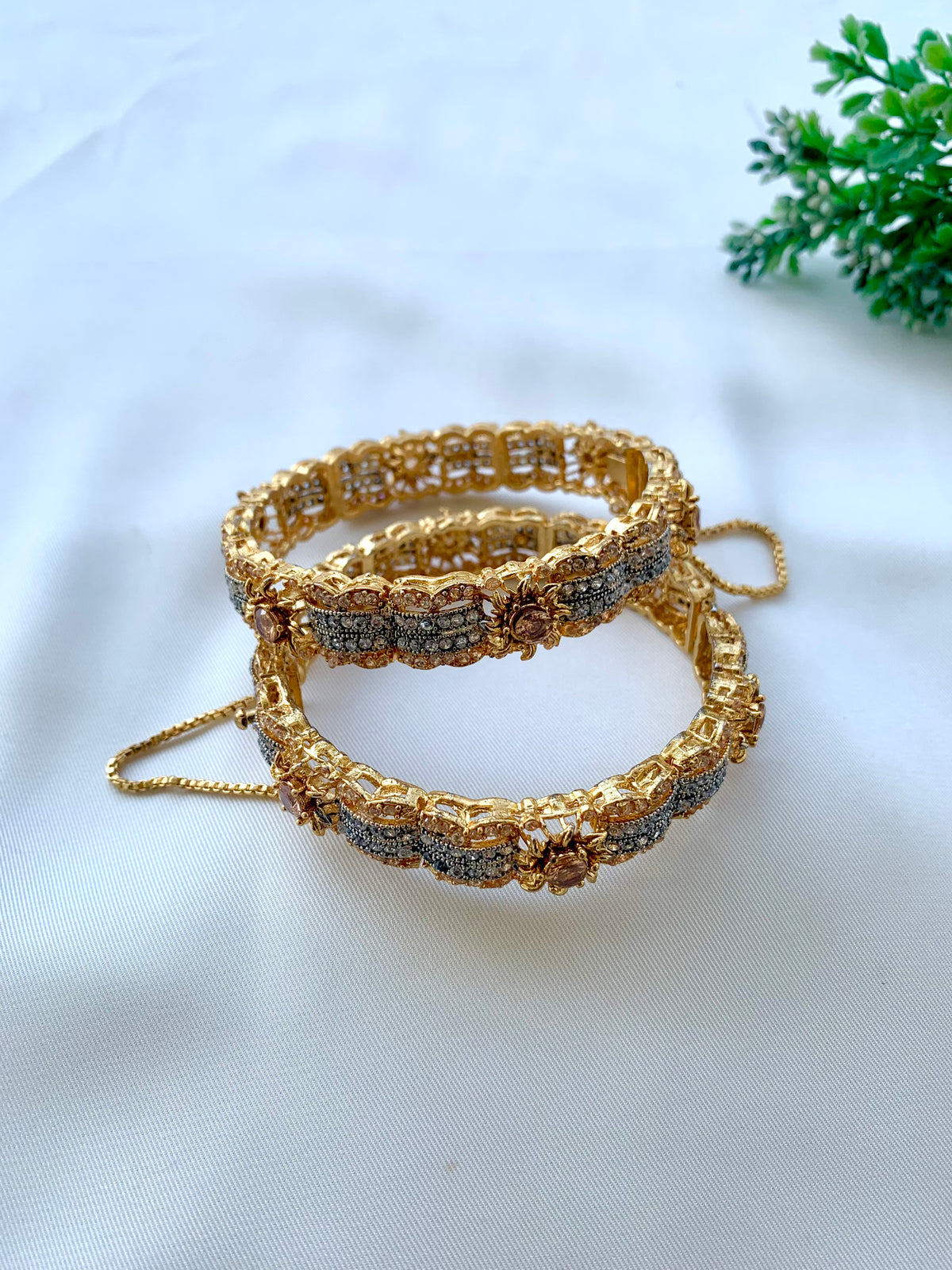 Two ornate gold-toned bangles with intricate floral detailing and small clear stones, displayed on a white fabric surface with a small green plant in the corner.