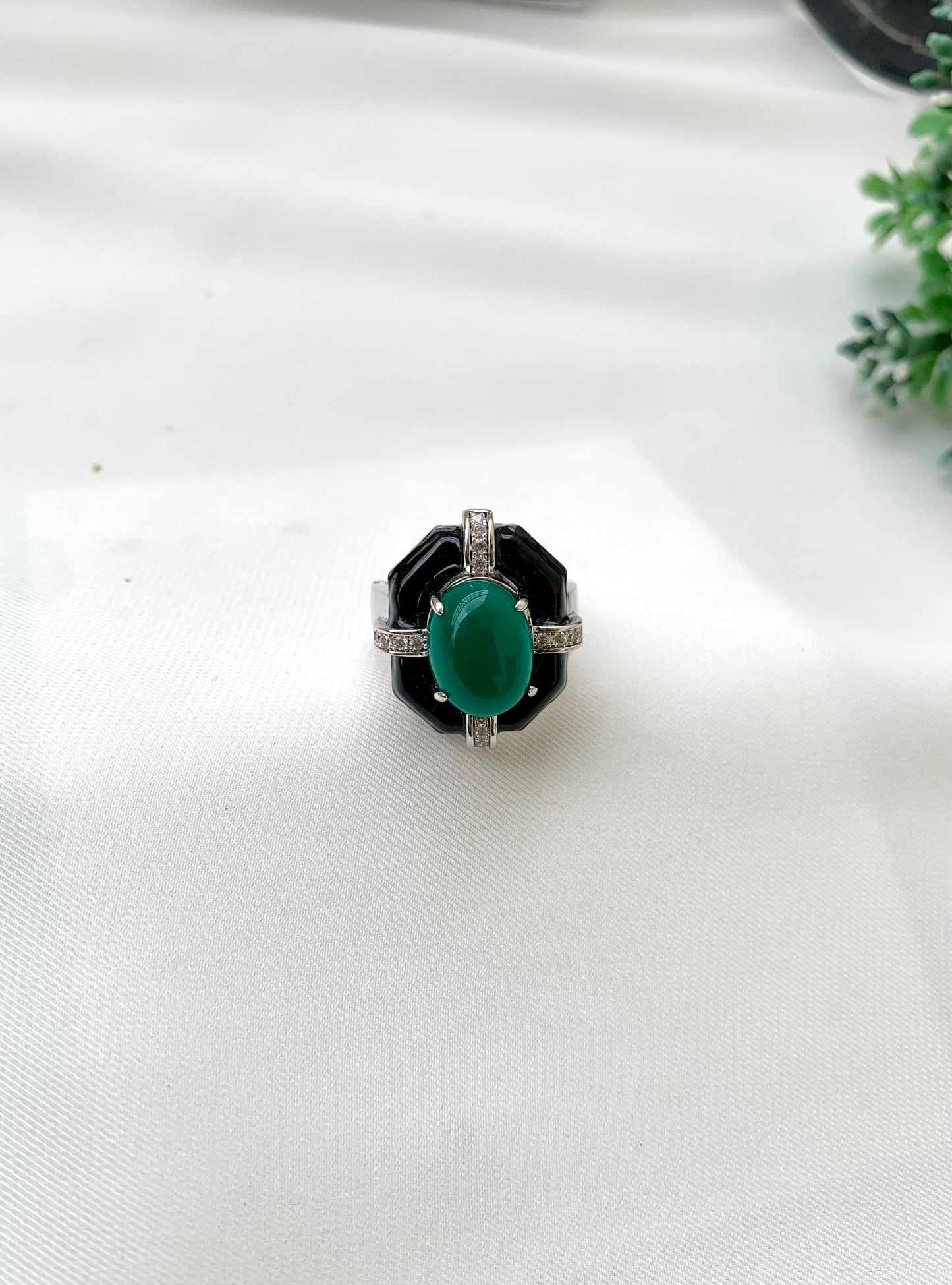 A vintage-style Art Deco ring featuring a central oval green stone, surrounded by a black inlay and a silver-toned band accented with small clear stones, resting on a white textured fabric surface.