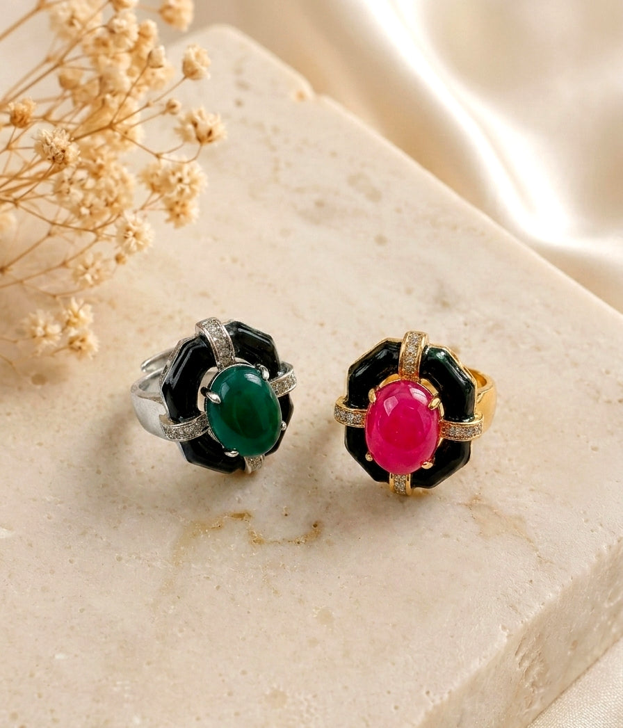 Two black enamel flower-shaped rings with oval center stones, one green and one pink, displayed on a beige stone slab with dried flowers and satin fabric.