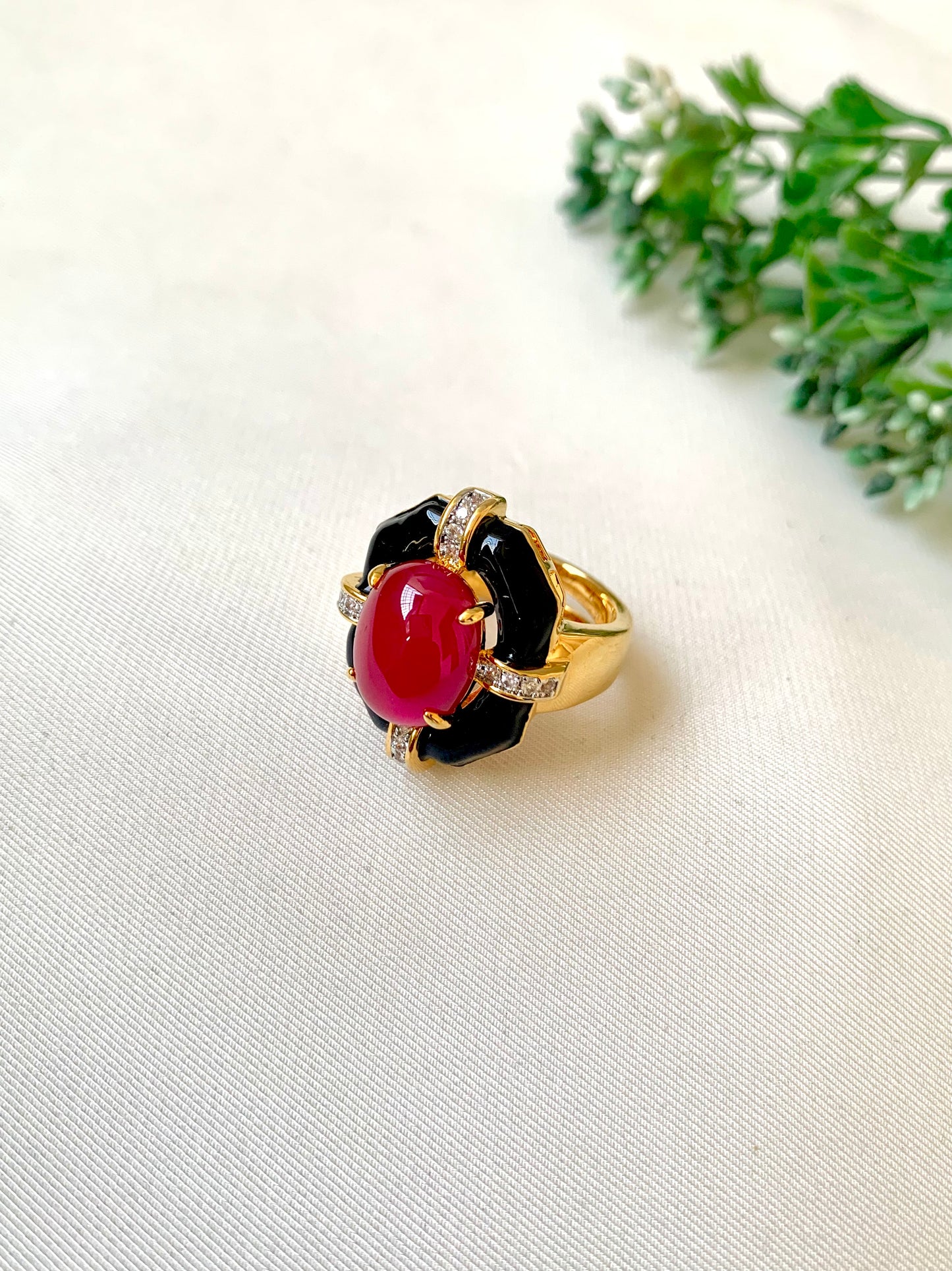 A gold ring with a black enamel floral setting and a prominent oval red cabochon gemstone, accented with small clear stones, resting on a white textured fabric with green leaves in the background.