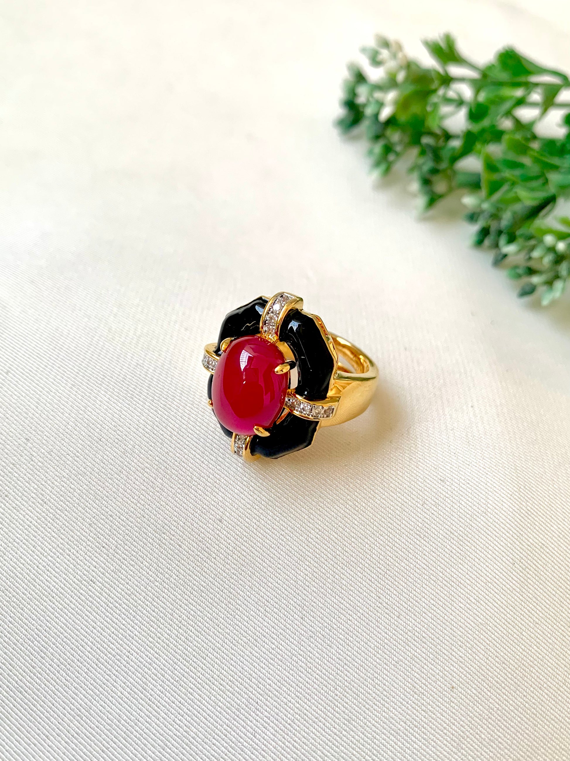 A gold ring with a black enamel floral setting and a prominent oval red cabochon gemstone, accented with small clear stones, resting on a white textured fabric with green leaves in the background.