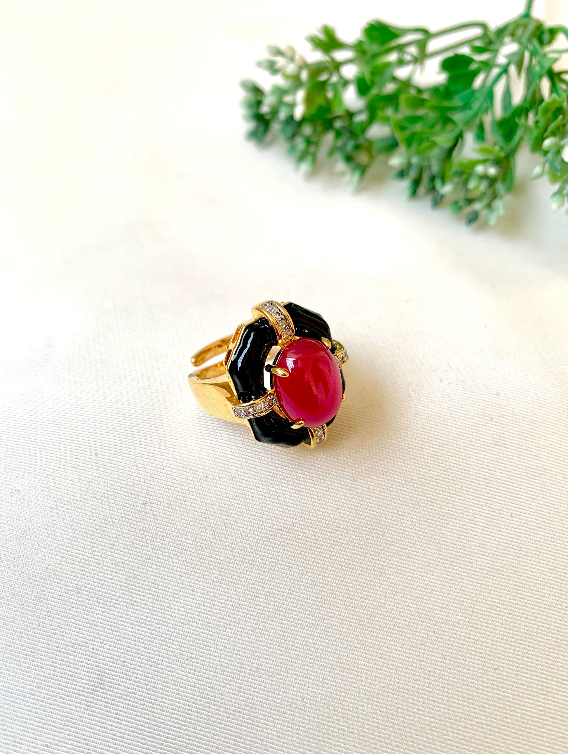 A gold ring with a large oval red cabochon stone and black stone accents, photographed on a light beige fabric surface with green leaves in the background.