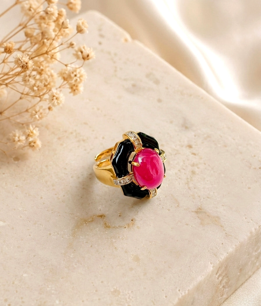 A gold ring with a black enamel flower-like setting and a prominent oval pink gemstone, displayed on a light marble block with dried flowers and satin fabric.