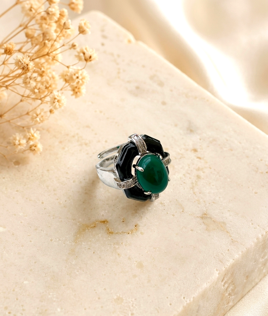 A silver ring with a green oval cabochon and black geometric side stones, displayed on a beige stone block with dried flowers.