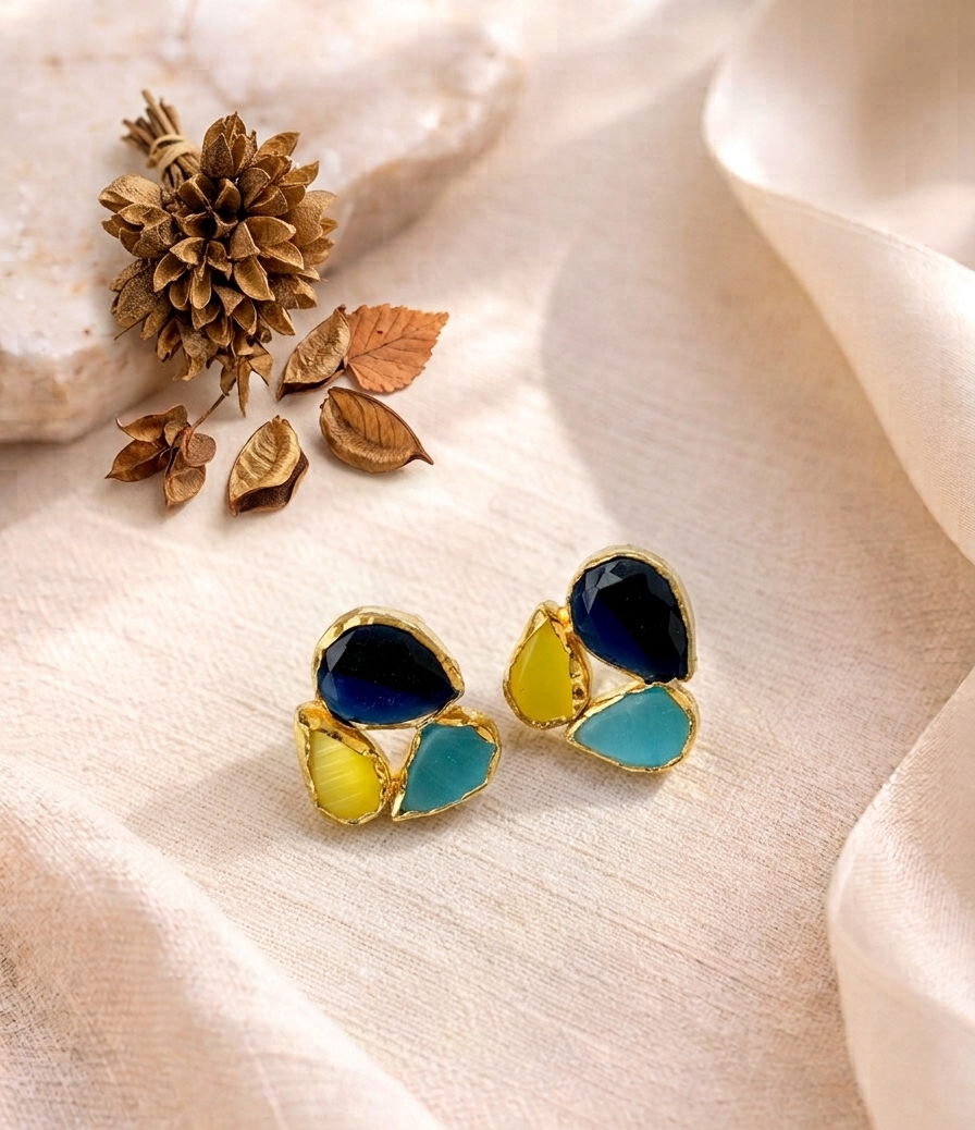 A pair of gold-tone stud earrings with layered teardrop-shaped enamel sections in deep navy blue, bright yellow, and light aqua blue, displayed on a beige fabric surface with dried botanical props.