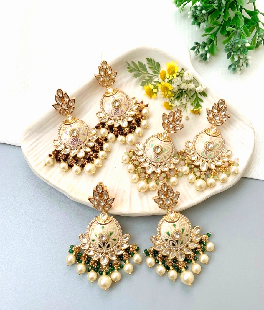 Four pairs of ornate gold-toned chandelier earrings with pearl-like beads, floral enameled details, and green accents, arranged on a white dish with small yellow flowers and greenery.