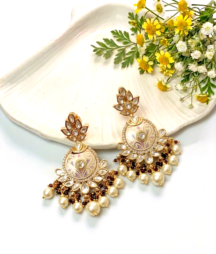 A pair of ornate gold-toned statement earrings with floral motifs, white pearl-like drops, and dark brown bead accents, styled on a white shell-shaped tray with small yellow and white flowers.