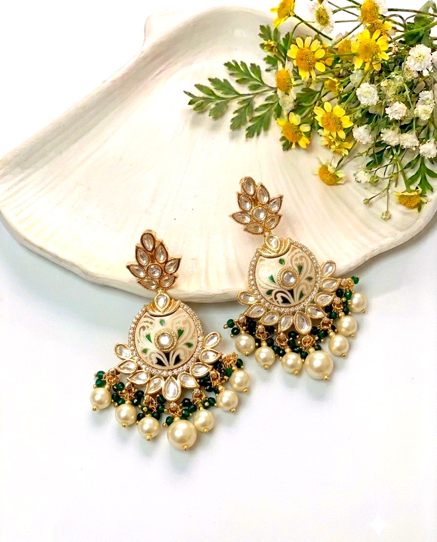 A pair of ornate gold-toned Indian jhumka earrings decorated with white faux pearls, green enamel, and clear rhinestones, displayed on a white scalloped tray next to yellow and white flowers.