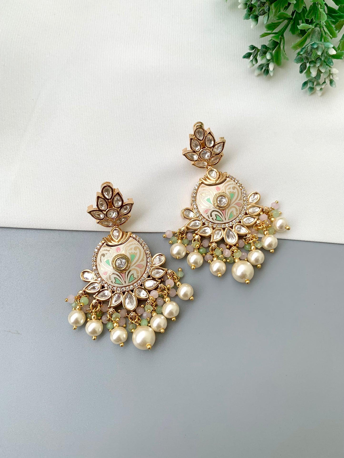 A pair of ornate gold-toned Indian jhumka earrings with floral enameled details, clear stone accents, and dangling pearl-like beads, displayed on a split white and gray surface with a small green plant in the corner.