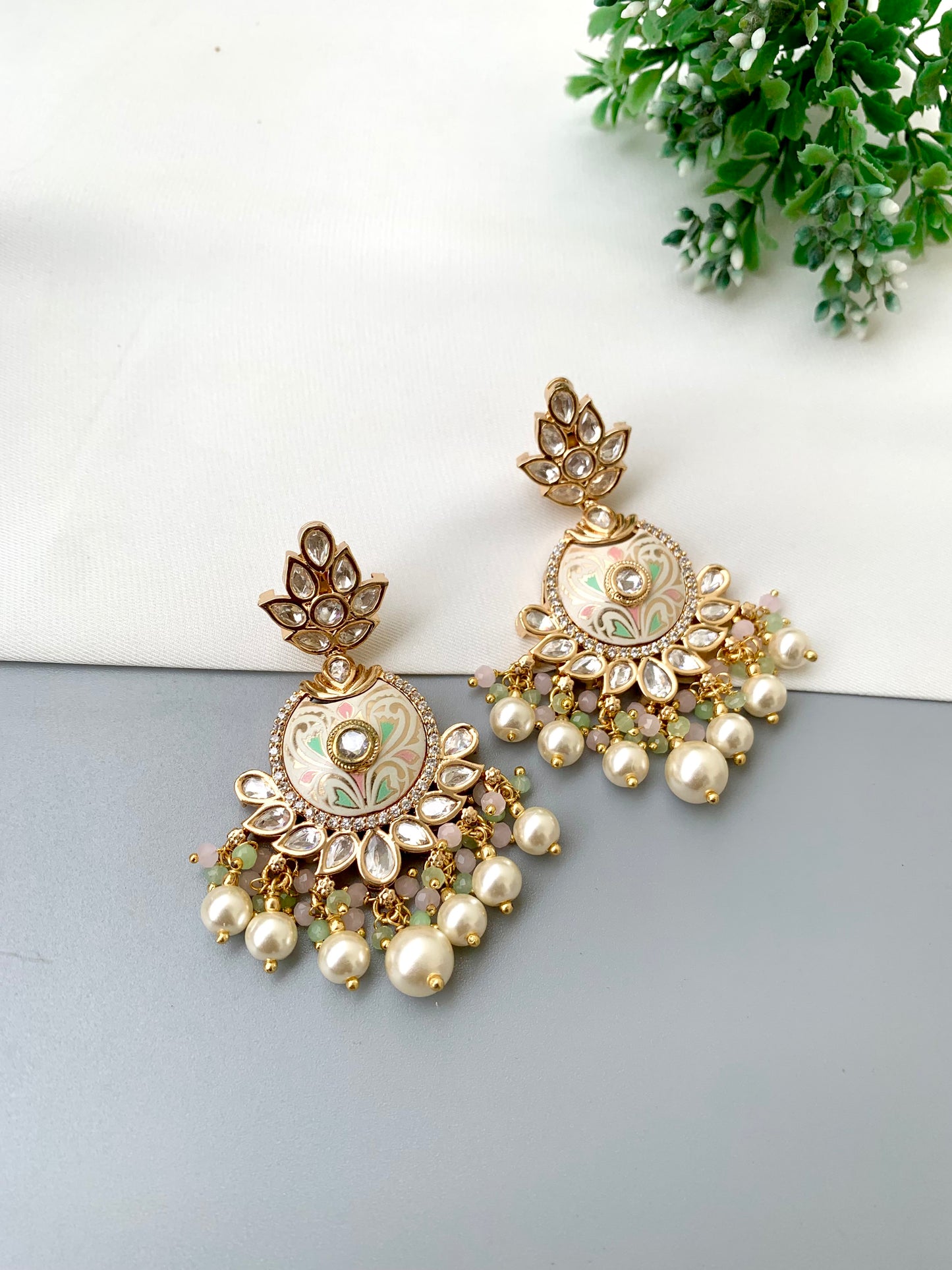 A pair of ornate gold-toned jhumka earrings with green and white enamel detailing, adorned with faux pearls and small stones, displayed on a split white and gray surface with a green plant in the corner.