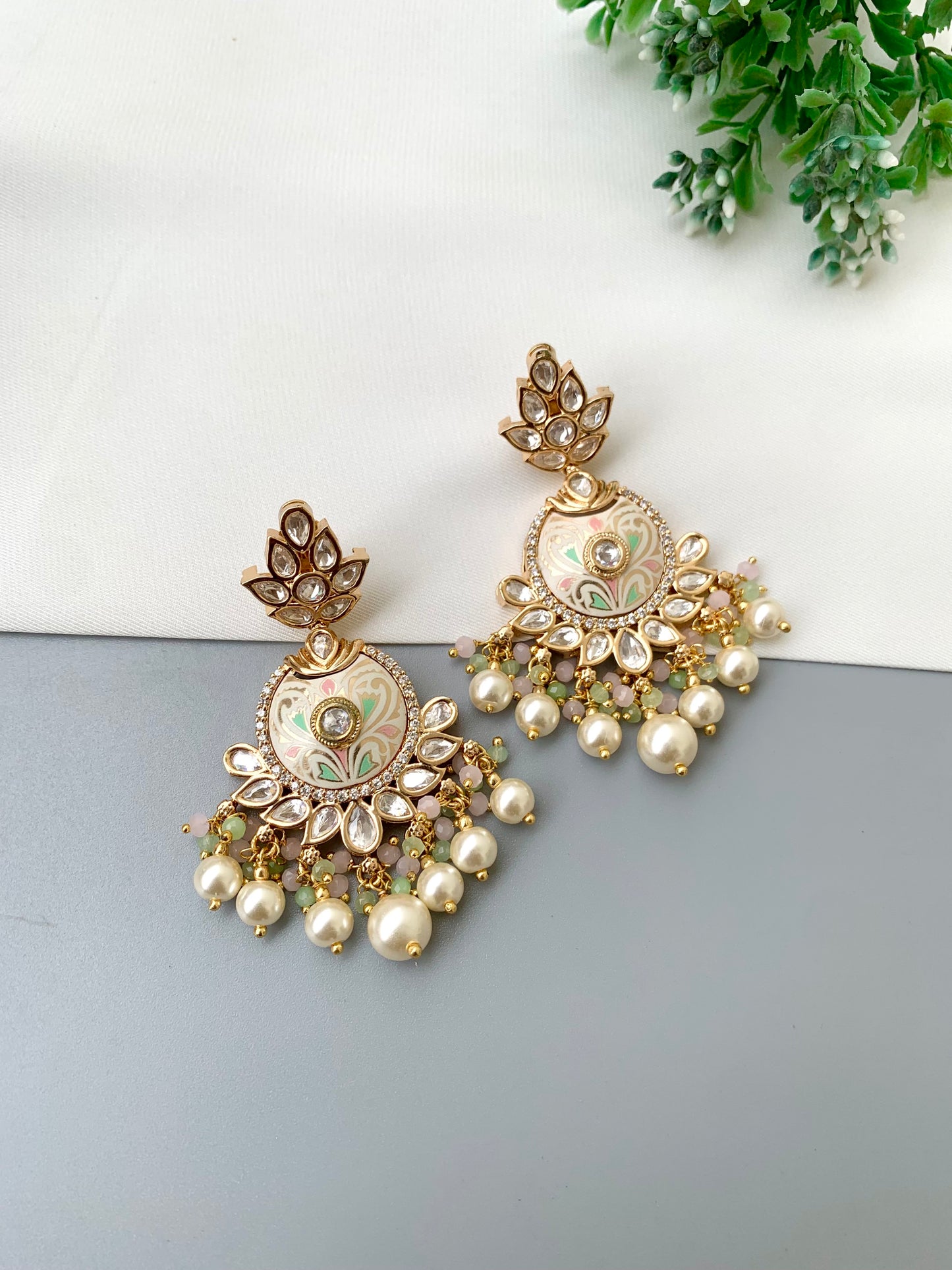 A pair of ornate gold-toned Indian jhumka earrings with green and white enamel detailing, clear stone embellishments, and dangling pearl-like beads, displayed on a split white and gray surface with a small green plant in the corner.