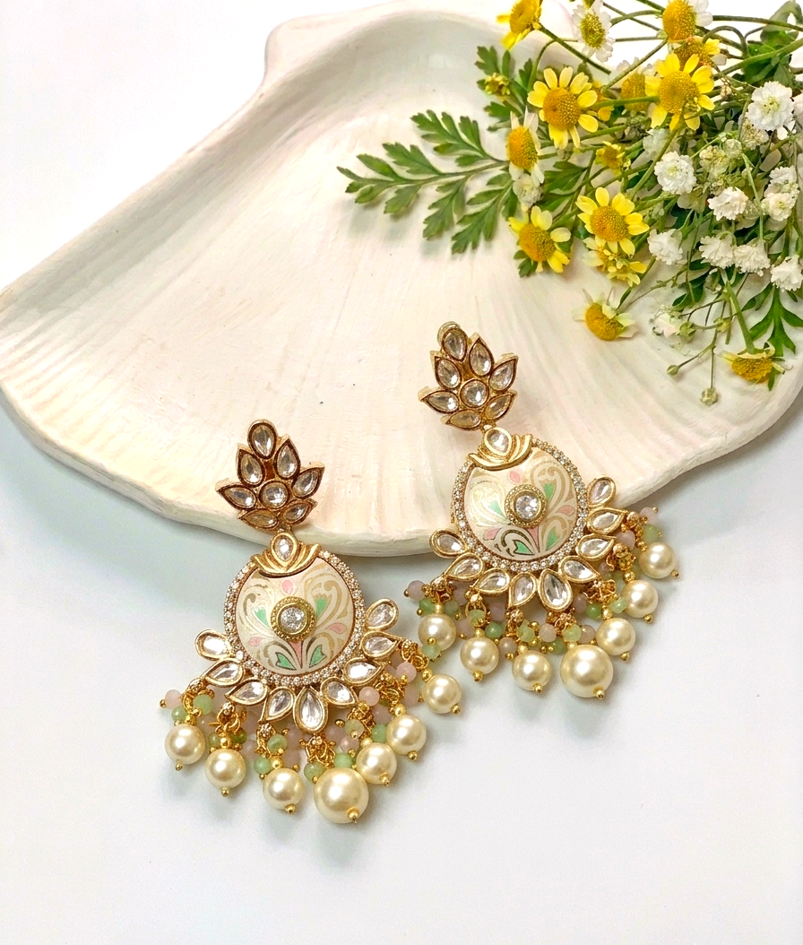 A pair of ornate gold-toned statement earrings with pearl-like drops and small clear stones, styled on a white shell with yellow and white flowers against a white background.
