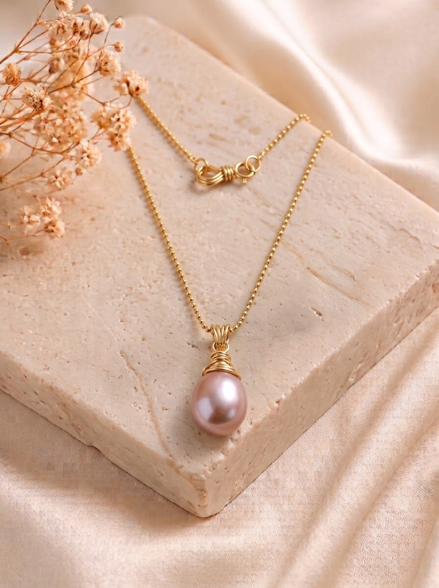 A gold chain necklace with a teardrop-shaped pink pearl pendant, styled on a textured beige stone block with dried flowers and draped satin fabric.