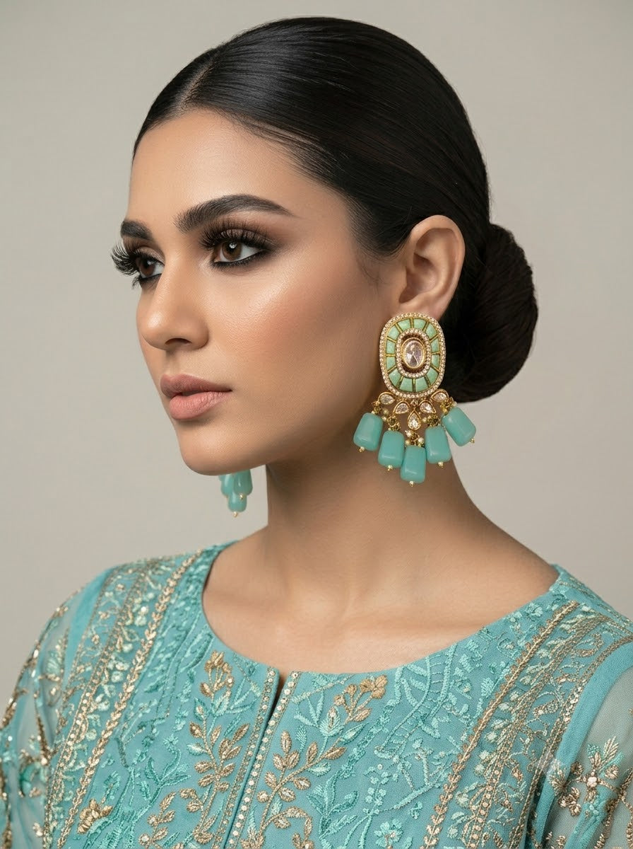 A woman in profile wearing a large ornate turquoise and gold statement earring, paired with a matching turquoise embroidered top, against a neutral beige background.