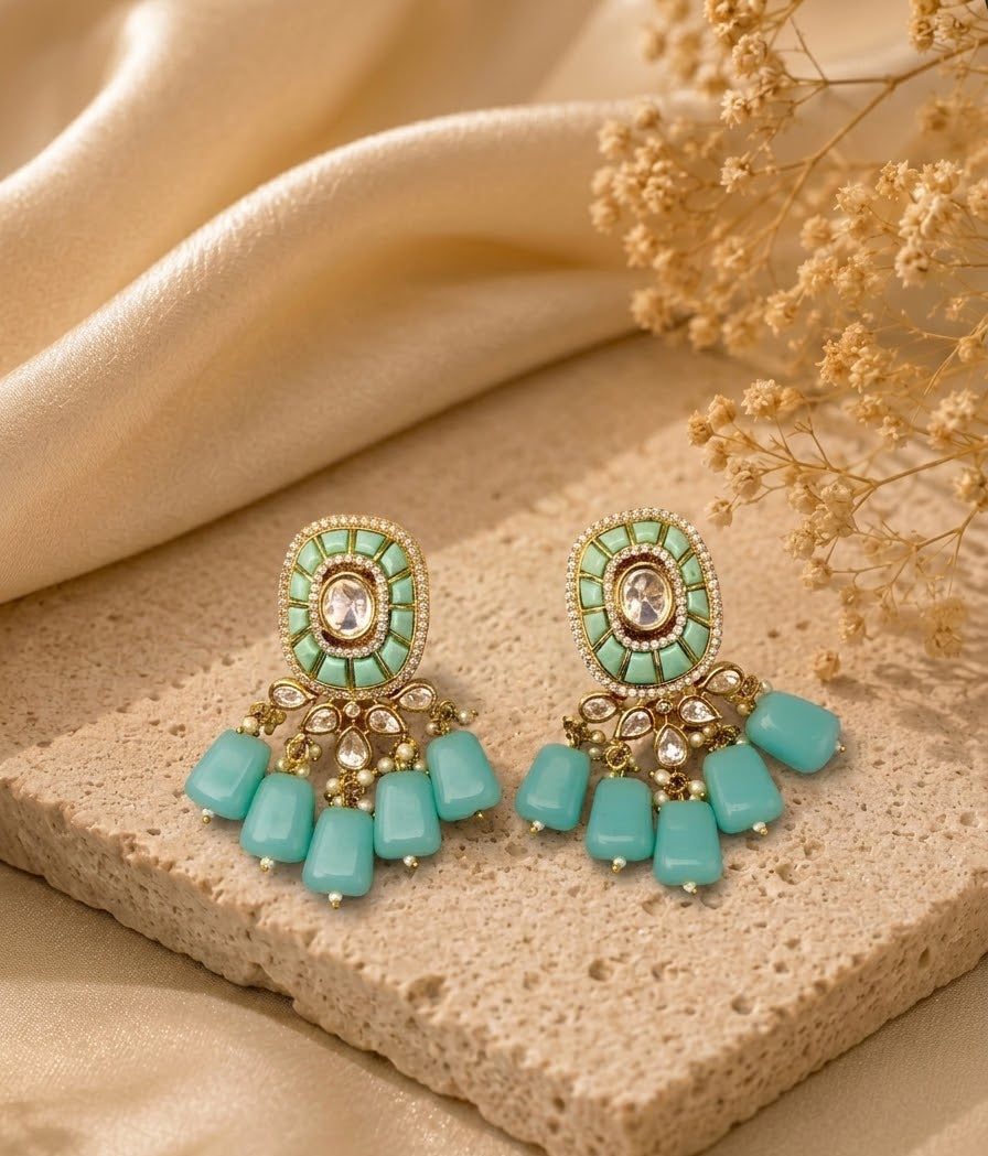 A pair of ornate gold-tone dangle earrings with mint green and turquoise-colored stones, displayed on a textured beige stone slab with draped cream fabric and dried flowers in the background.