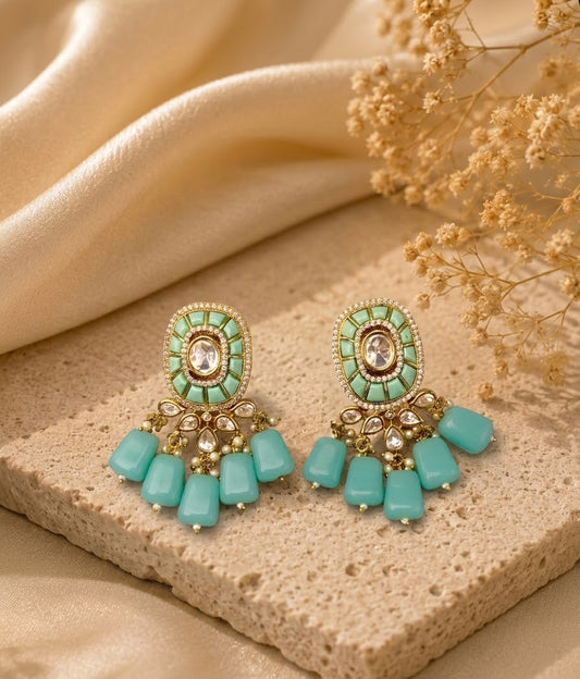 A pair of ornate gold-tone dangle earrings with mint green and turquoise-colored stones, displayed on a textured beige stone slab with draped cream fabric and dried flowers in the background.