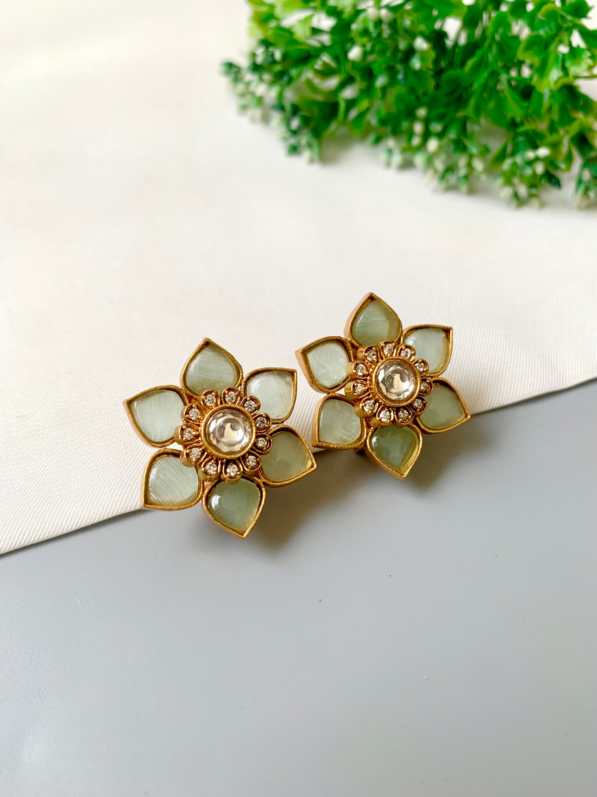 Floral earrings with gold and green accents on a white surface with greenery in the background