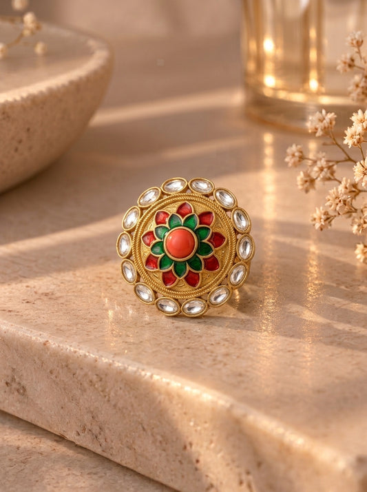 A gold-toned traditional Indian Kundan ring with a central red and green floral motif and clear stone accents, resting on a beige stone surface with warm, soft-focus background props.