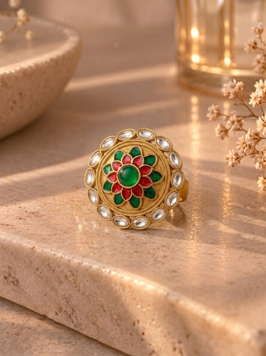 A gold-toned mandarin ring with a green and red floral center and clear stone accents, resting on a beige stone surface in warm sunlight.