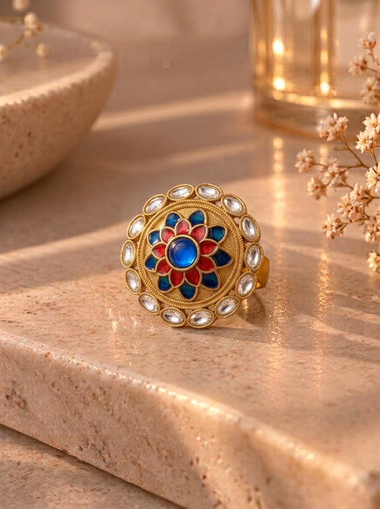 A gold-toned mandarin-style ring with a central blue stone and red, blue, and gold enamel floral pattern, accented with small clear stones, resting on a beige stone surface.