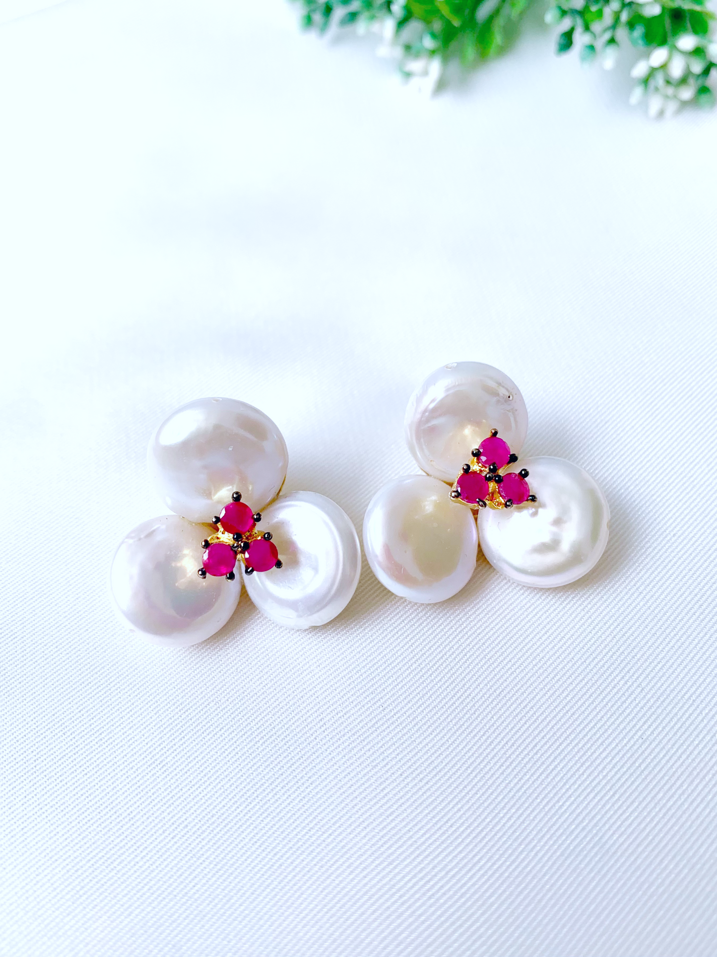 Three white baroque freshwater pearls arranged in a cluster with a small central floral accent featuring bright pink gemstones and gold metal details, displayed on a white textured fabric background.