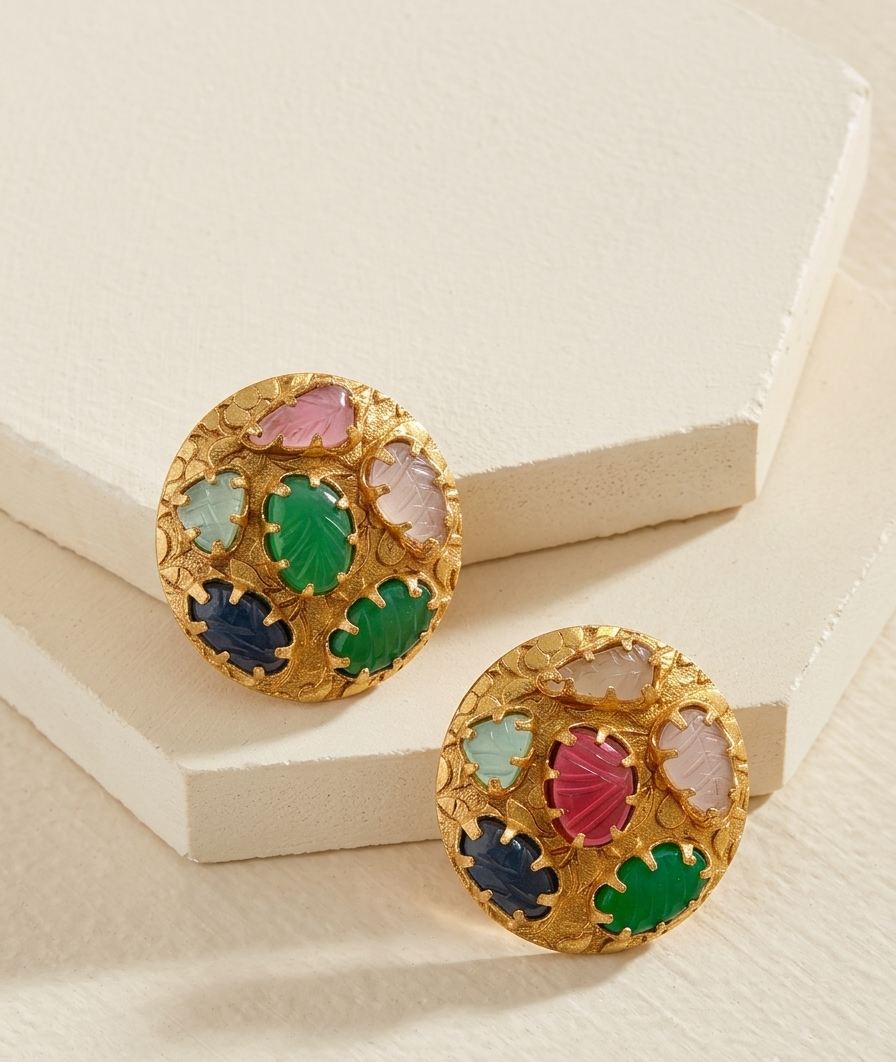 A pair of round gold-toned earrings with colorful inlaid shell-like pieces in pink, green, blue, and white, displayed on a cream-colored geometric surface.