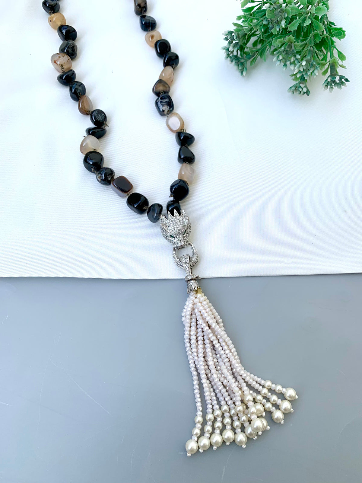 A beaded necklace with black and brown stone beads, a silver-toned lion head clasp, and a long pearl tassel, displayed on a split white and gray background with a small green plant in the corner.