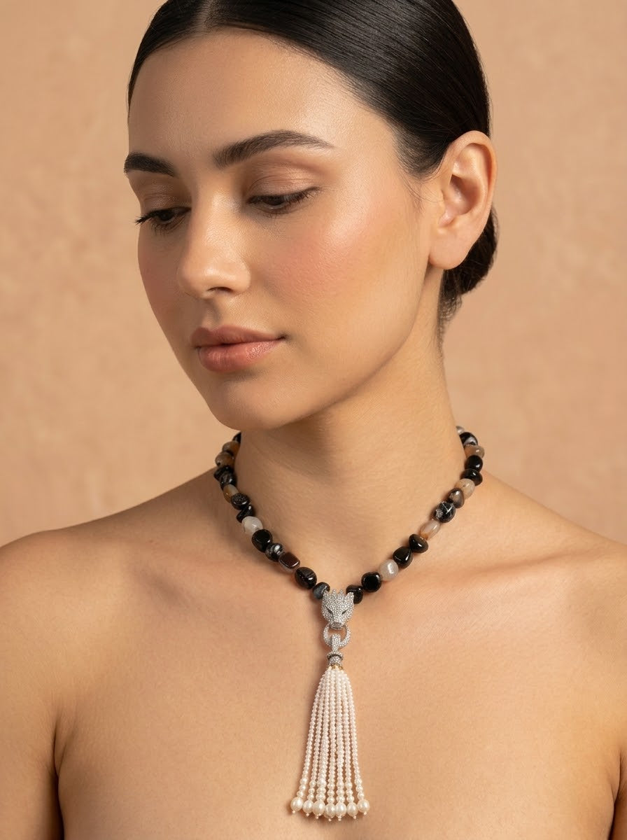 A woman wearing a beaded necklace with black and white beads, a silver-toned central connector, and a long white tassel pendant, photographed against a warm beige studio background.