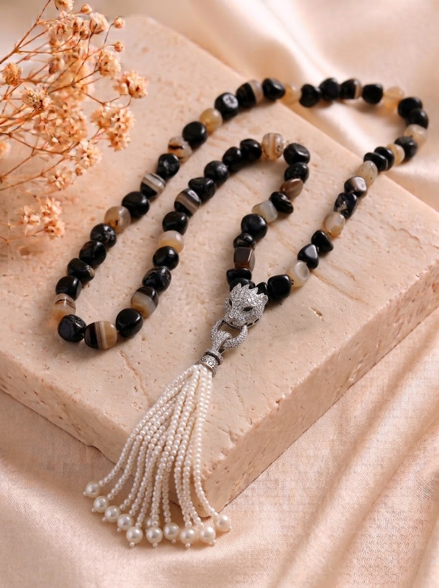 A beaded necklace with black and brown agate-like beads, a silver-toned lion head pendant, and a long white pearl tassel, displayed on a beige stone block with dried flowers.