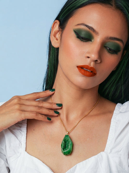 A close-up lifestyle shot of a model wearing a gold chain necklace with a green teardrop-shaped agate pendant. The model has dark green hair, green eyeshadow, orange lipstick, and is wearing a white top against a light blue background.