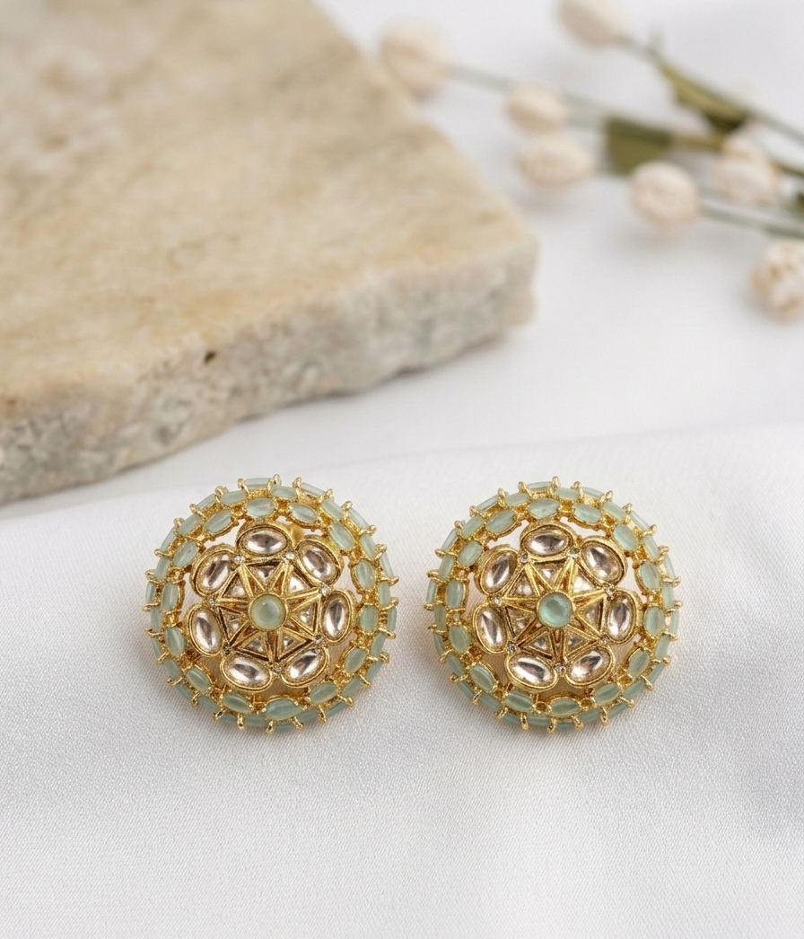 A pair of gold-plated mandarin earrings with light green stones and clear teardrop-shaped crystals, displayed on a white fabric surface next to a stone prop.