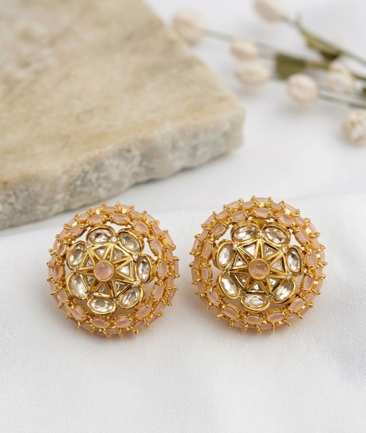 A pair of gold-toned round stud earrings decorated with clear crystals and small pale pink stones, displayed on a white surface with a beige stone slab and dried flowers in the background.