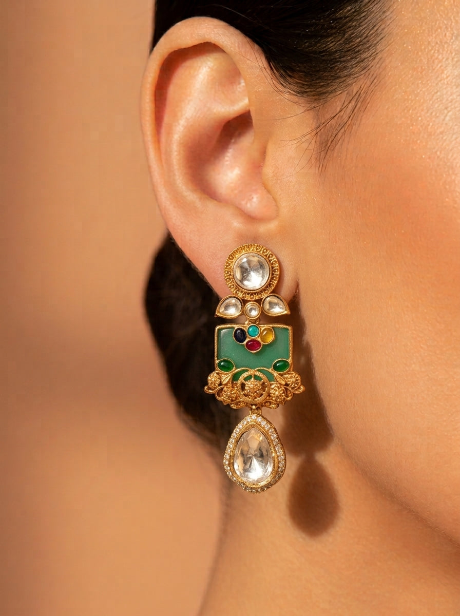 A close-up of a woman's ear wearing a detailed gold-toned Indian-style dangle earring with a green enamel panel, multicolor accents, and clear stones.