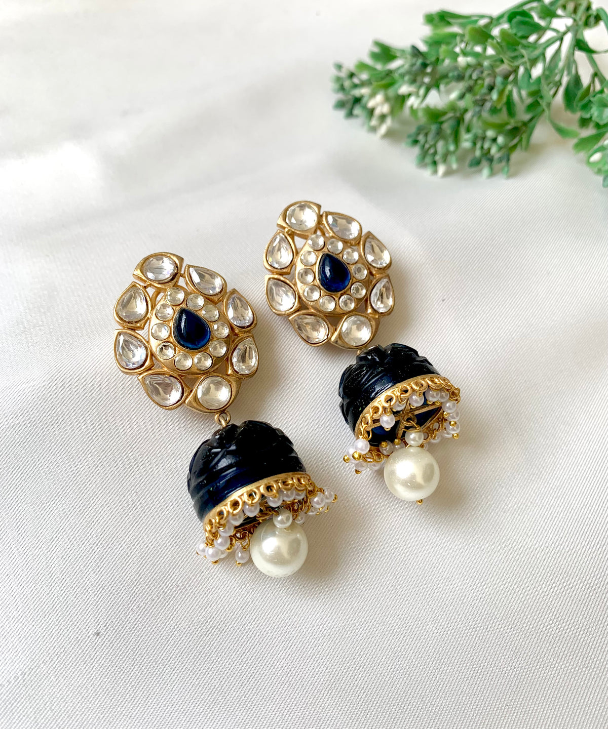 A pair of gold-plated earrings adorned with polki kundan, semi-precious stones, and pearls, displayed against a white background with a green leafy element.