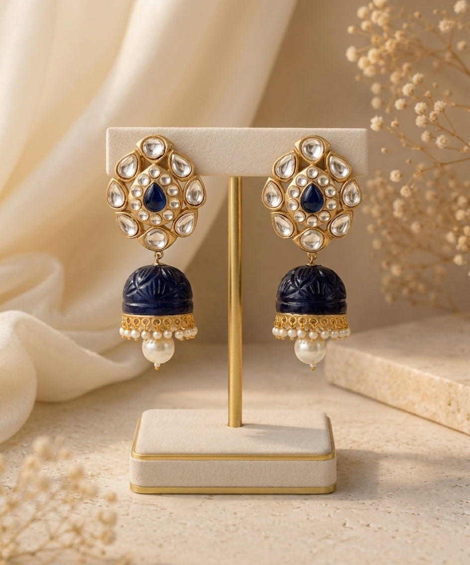 A pair of ornate gold-toned earrings displayed on a cream jewelry stand. Each earring features a floral-shaped gold setting with clear stones, a deep blue teardrop center, and a dangling deep blue bell-shaped charm with gold detailing and a small pearl at the bottom.