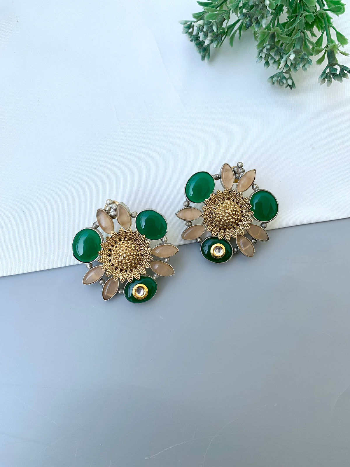A pair of gold-toned flower-shaped earrings with green oval stones, clear teardrop rhinestones, and small dangling green beads, displayed on a light gray surface with a white background and green leaves in the corner.