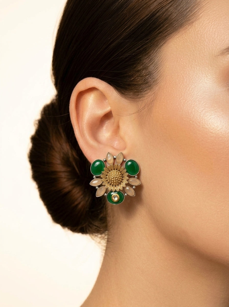 A close-up of a model's ear wearing a gold-toned flower-shaped earring with green oval stones and clear rhinestone petals, against a plain light background.