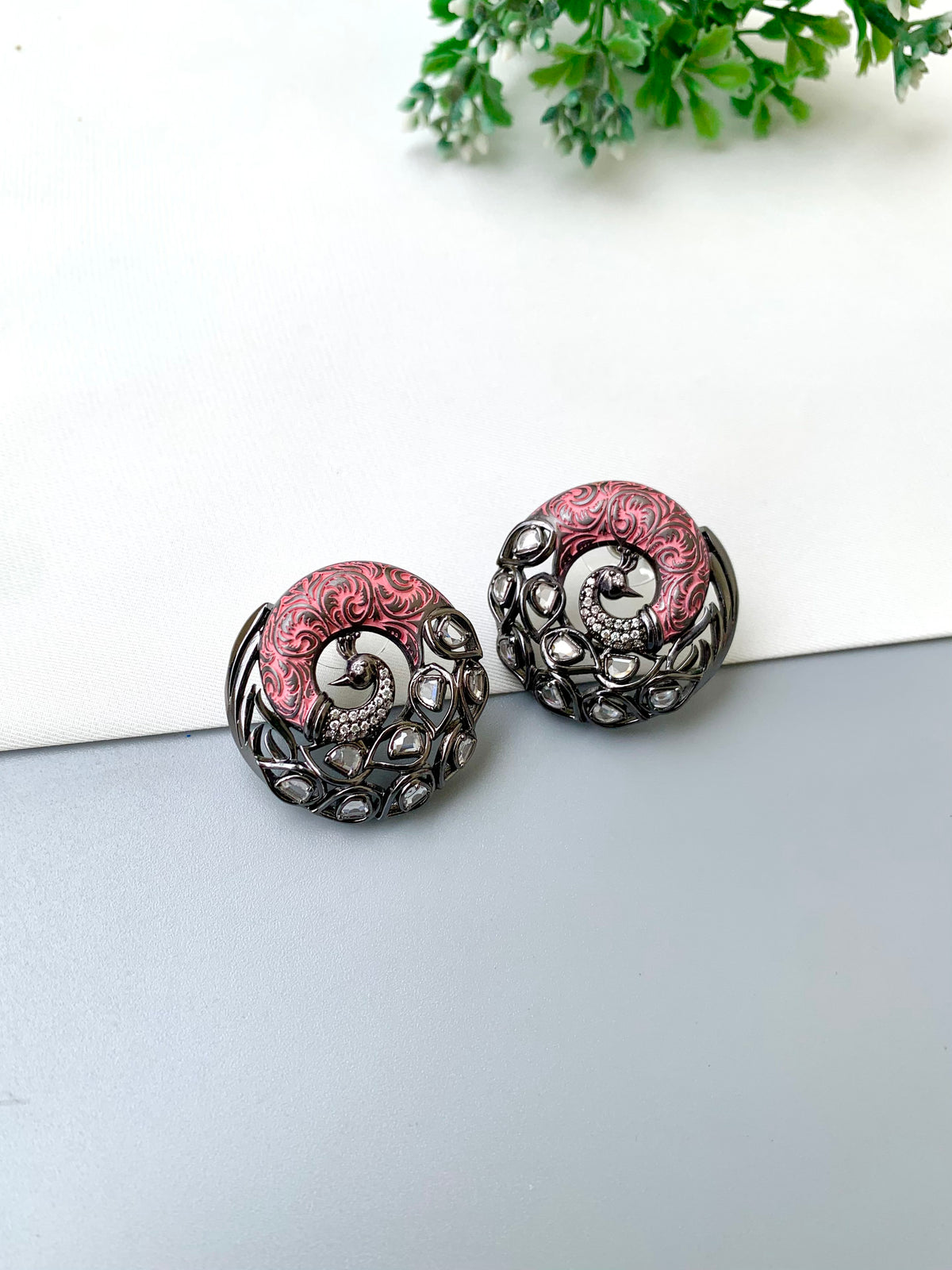 A pair of round, ornate earrings with a silver-toned filigree border and a central pink enamel spiral motif, displayed on a split white and gray surface with a small green plant in the background.