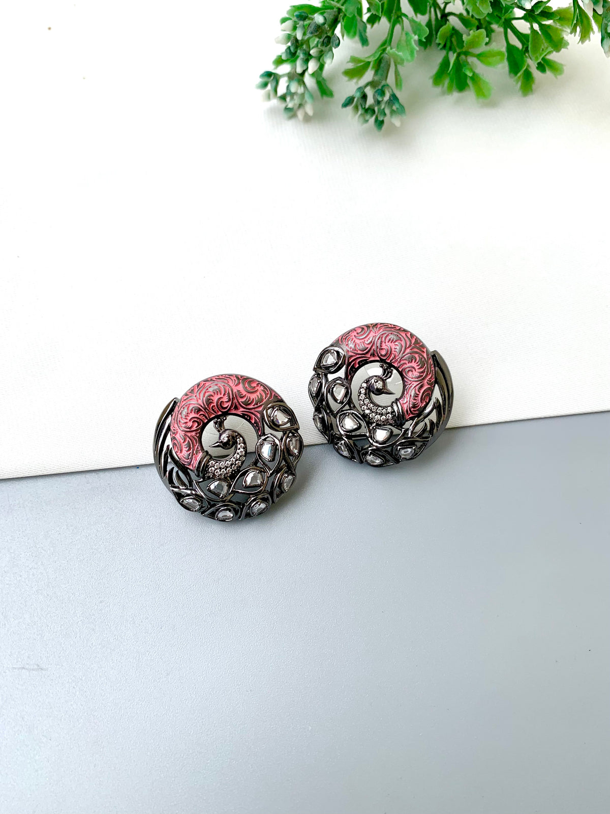 A pair of round, ornate earrings with a black and silver filigree design and pink floral accents, displayed on a split white and gray surface with a small green plant in the background.