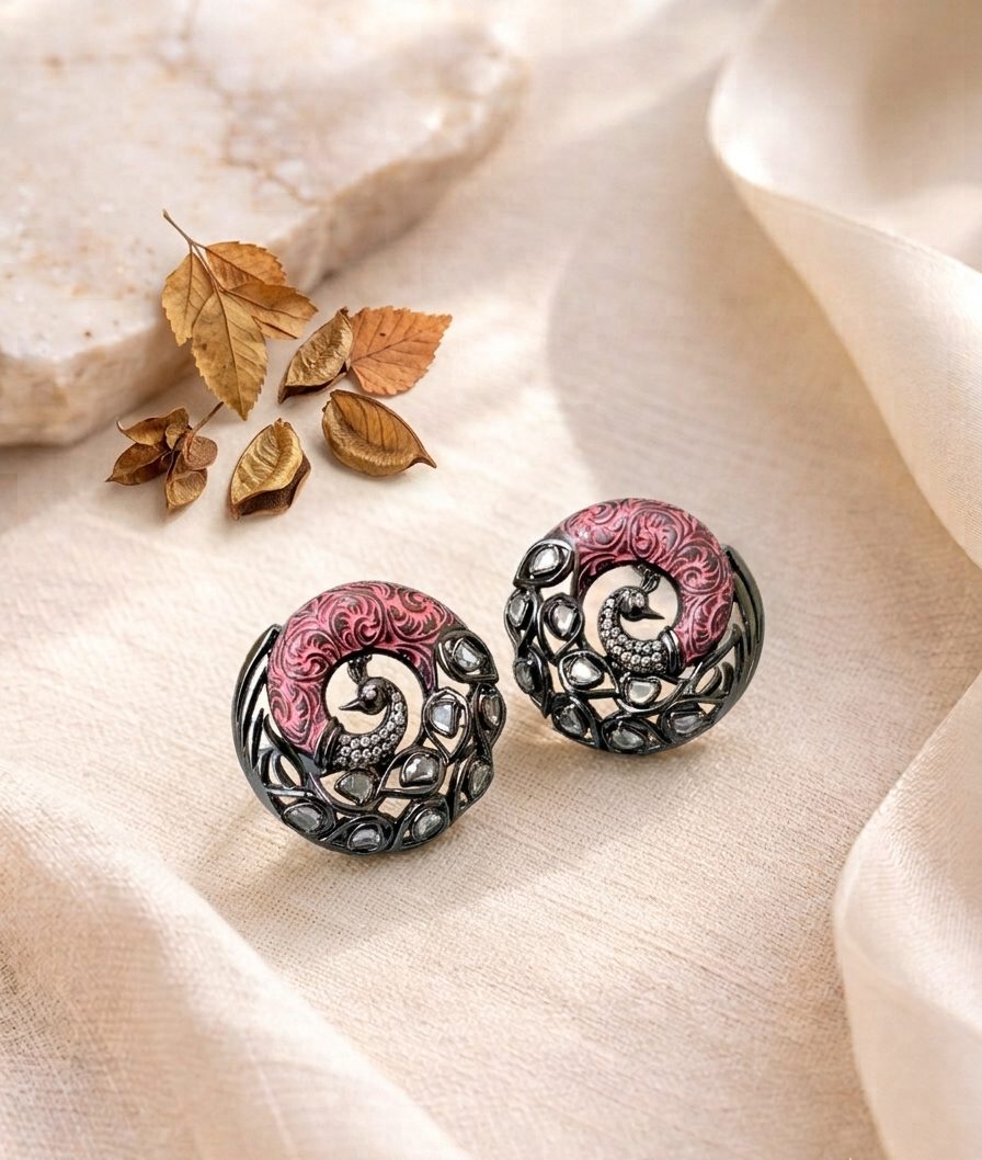 A pair of round statement earrings with intricate filigree detailing, featuring a pink and silver spiral design, displayed on a beige fabric background with dried leaves and a stone prop.