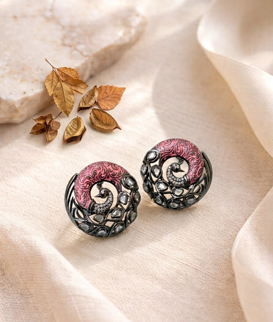 A pair of round statement earrings with intricate filigree detailing, featuring a pink and silver spiral design, displayed on a beige fabric background with dried leaves and a stone prop.